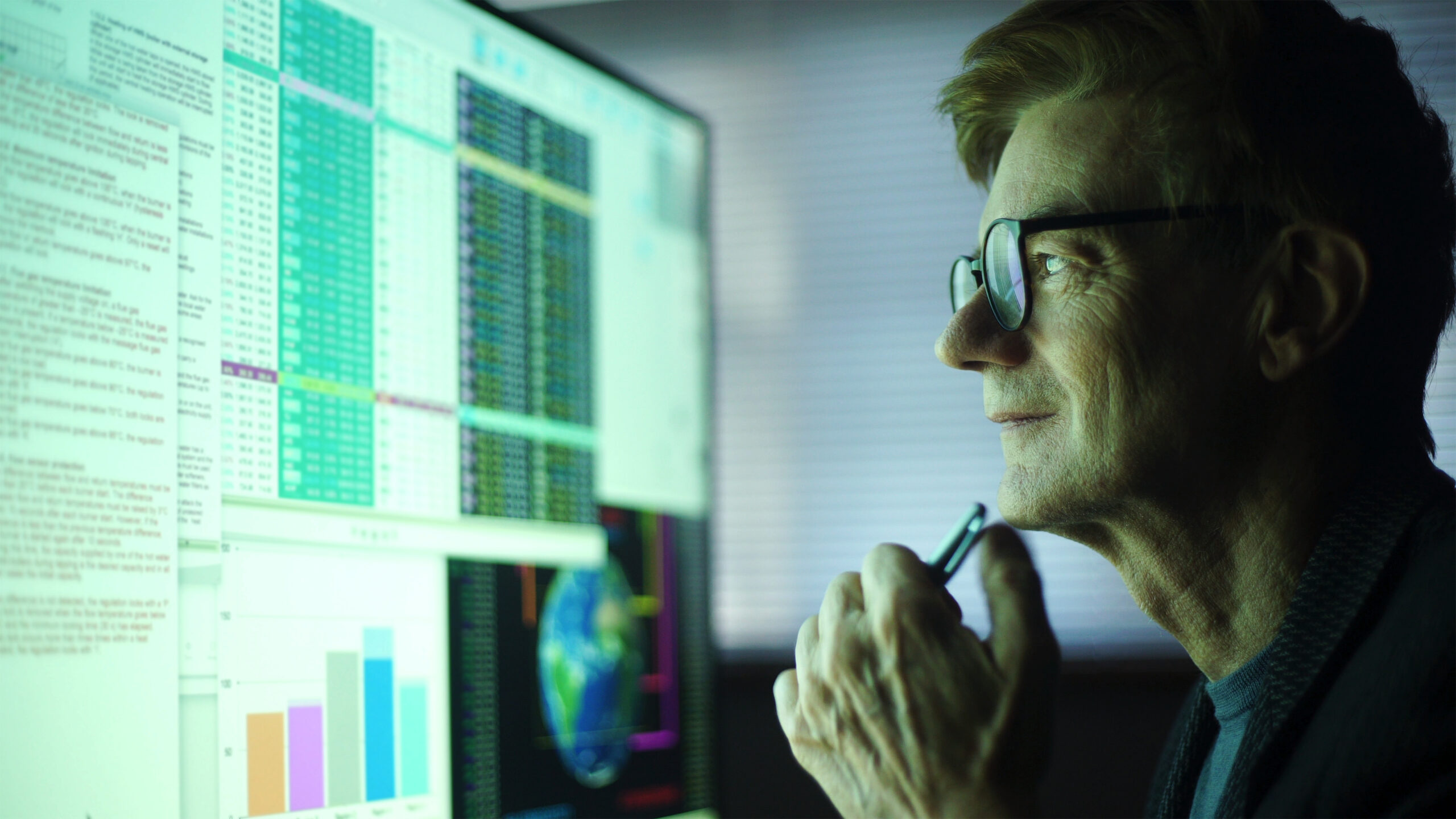 Stock photo of a mature man working in his home office. He’s studying a large computer monitor displaying a variety of numerical data, global information & text.